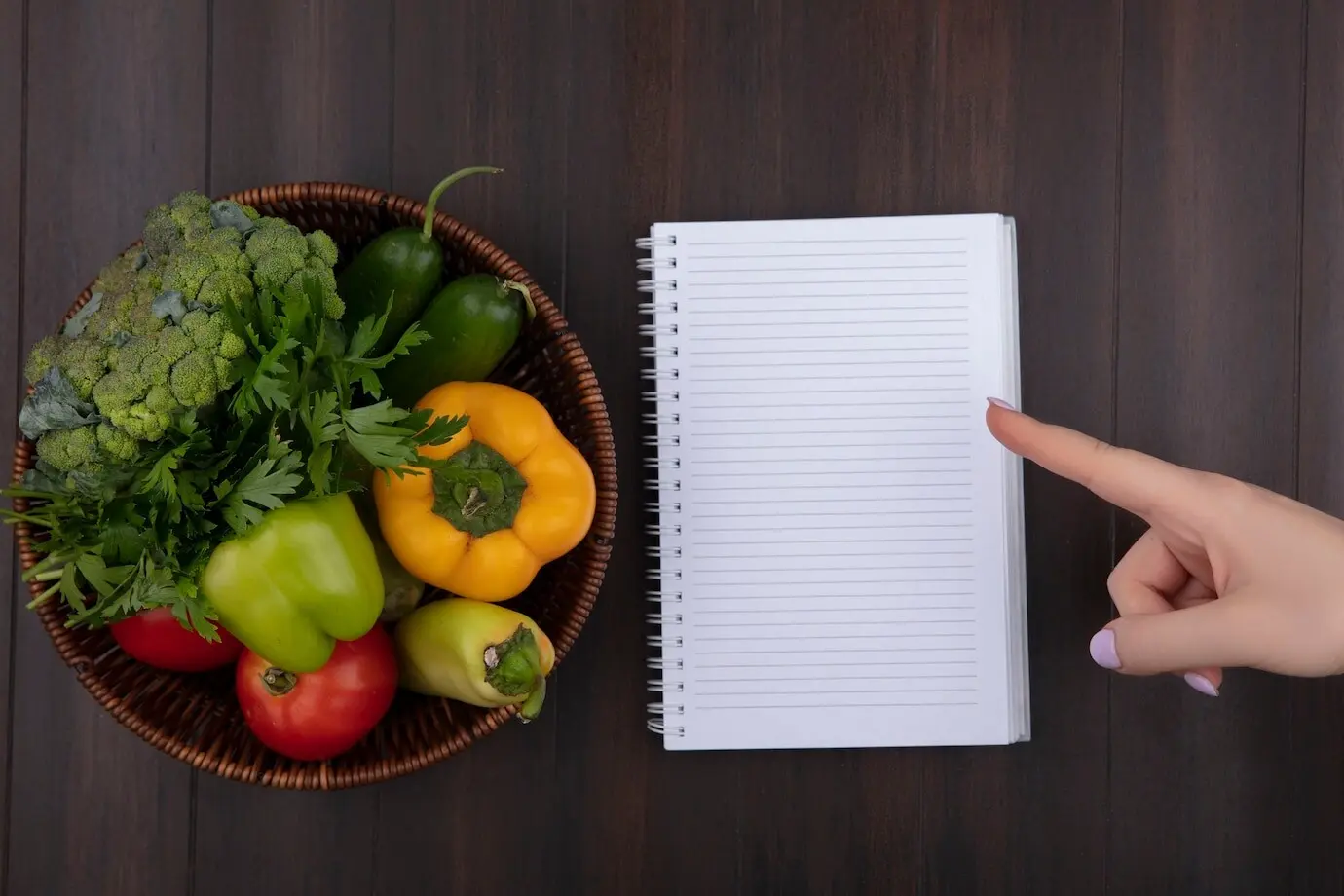 Ansicht von oben mit Platz für Text; eine Frau zeigt auf ein Notizbuch mit Paprika, Petersilie und Tomaten in einem Korb auf Holzuntergrund.