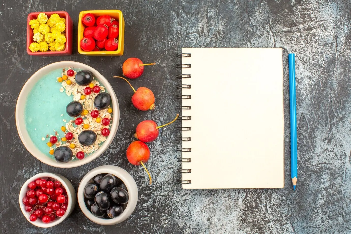 Nahaufnahme von oben mit Beeren, Notizbuch, Bleistift, Kirsche, bunten Beeren und Haferflocken in den Schalen.